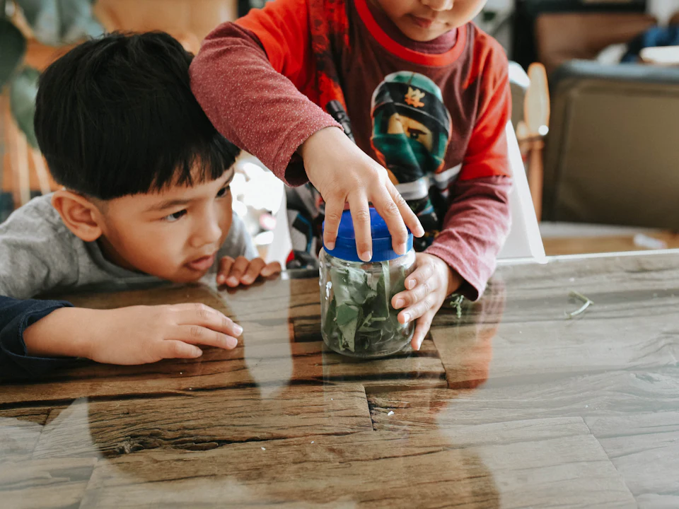 Dos niños guardan dinero en un frasco sobre una mesa de madera.